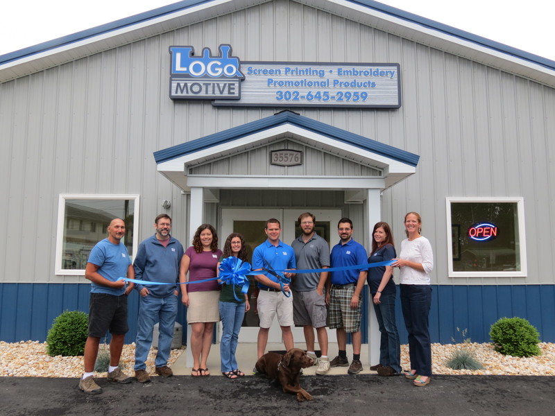 The team at Logo Motive Custom Apparel, including (l-r) Mike Kakas, Mike Smith, Crystal Heidel, Candace Rogers, owner Billy Hein, Alan Felts, Mark Hayduk and Allison Thomas join Meghan McCalley of Rehoboth Beach-Dewey Beach Chamber of Commerce and Duke, center, head of public relations and security, for a grand opening celebration. SOURCE SUBMITTED