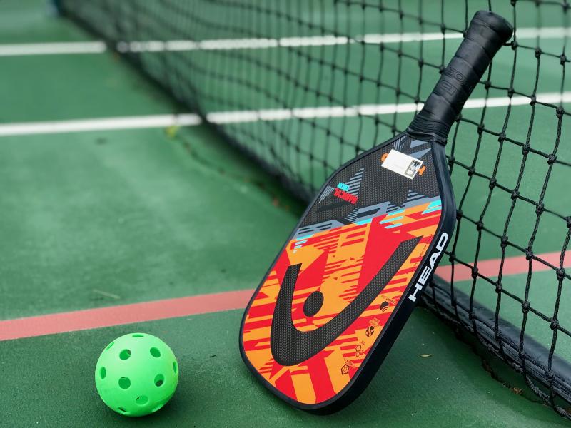 A pickleball, a pickleball paddle and the red boundary lines of a pickleball court overlaid on a tennis court at the Canalfront Park courts in Lewes. The pickleball is like a Wiffle ball but a little larger, a little heavier, and with round holes vs. oblong holes on a Wiffle ball. The holes slow the progress of the ball through the air, giving the pickleball a unique feel on the court. DENNIS FORNEY PHOTO