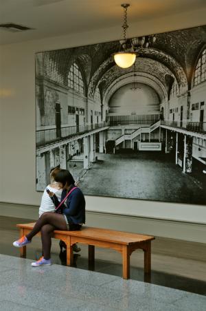In a clash of eras, two visitors to the Ellis Island museum seem oblivious to the large black and white photograph of the Registry Room behind them. BY RON MACARTHUR