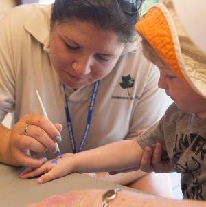 Sarah Schapira, a naturalist intern gives Aidan Healy, 2, a shark tattoo at the face-painting table. BY DENY HOWETH