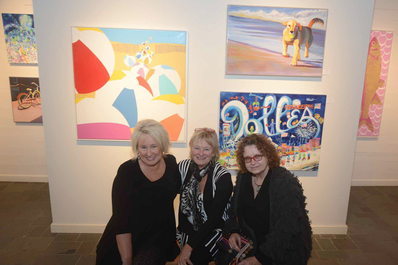 In the Tubbs Gallery, artists (l-r) Nina Mickelsen, Tara Funk Grimm and Jan Crooker sit in front of images of their collective work. The image on the left is Mickelsen’s “Can’t Wait to Get To The Beach.” The image on top right is Crooker’s “Beach Dog.” On the bottom right is Funk Grimm’s “4th of July.”