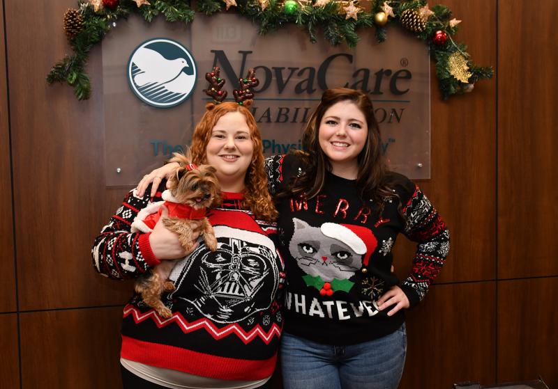 Showing off their “ugly” Christmas sweaters are NovaCare Rehabilitation employees Kristin Phillips, left, and Bridget Windsor and Maddie the dog.