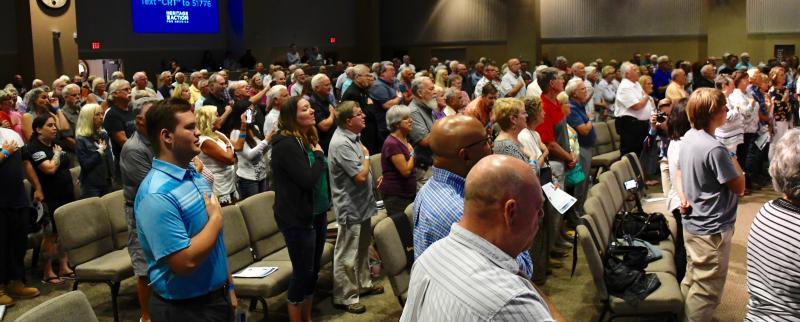 A large crowd at Crossroad Community Church stands to say the Pledge of Allegiance prior to the start of the forum.