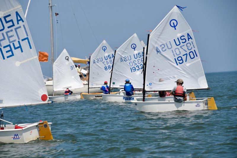 Optimist red, white and blue fleet sailors jockey for position at the starting line with the race committee boat in the background.