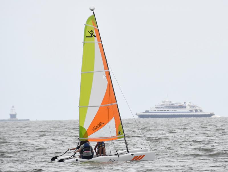 With two Lewes icons in the background – Harbor of Refuge Lighthouse and Cape May-Lewes Ferry – YMCA Explorers sail on a Hobie Cat.