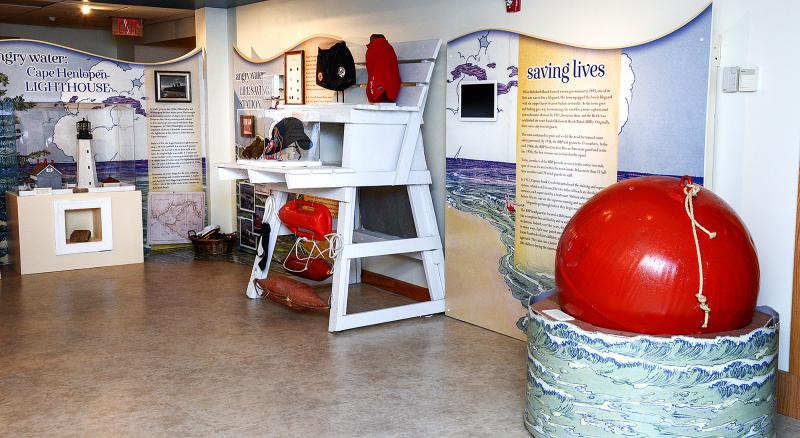 Red bouy on right side, lifeguard stand in the middle with varying equipment over time displayed, on left side in the back is a lighthouse station