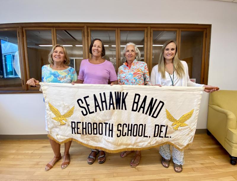 Holding the Rehoboth High School marching band banner in this photo from August 2022 are Rehoboth High alumni (l-r) Gail Nowakowski, Jackie Johnson Shockley and Hope Wilson Lavachia, with Cape Henlopen School District Public Relations Coordinator Steph DeMalto, who helped work to secure artifacts for a history room located in Rehoboth Elementary that will be open to the public Friday, Sept. 22. ELLEN DRISCOLL PHOTO