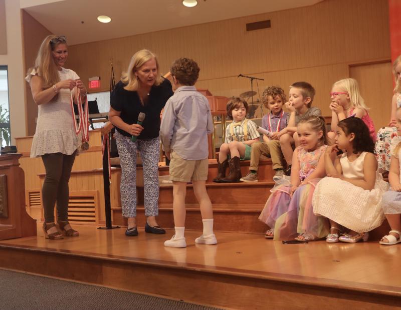 Director Betsy Plowman hands Owl Class graduate Oakes Thompson his diploma. Teacher Heather Mento eagerly waits to give him a medal. His excited fellow Owl Class graduates watch the exchange. In back are (l-r) Thorton Plummer, Teddy Parsons, Brixton Fehrenbach and Kate Creed. In front are Hanna Mae Baker and Camila Morales.