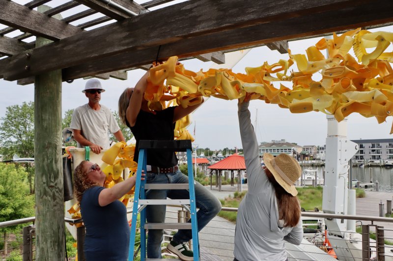 It took a team – and a Board of Public Works bucket truck – to hang Niki Lederer’s art on the trellises in Canalfront Park. Shown are (l-r) Mark Wheatley, Lewes Board of Public Works; Giulia Kirsch, Lewes Parks and Marina administrator; Heidi Lowe, Lewes Public Art Committee chair; and Niki Lederer, artist.