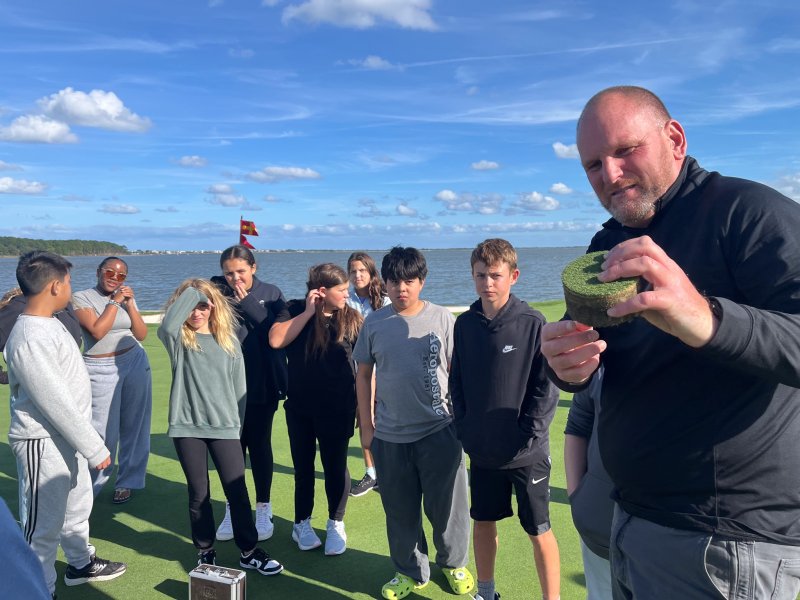Alan Fitzgerald from First Green, right, shows Beacon students (l-r) Mateo Tupaz, Portia Bowden, Briah McCabe, Juliana Lebron, Gabriella Brittingham, Charlotte Marchetti, Roman Ramirez and Lucan Skonieczki the plug from a golf course hole.