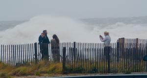 Huge waves break behind people as they take photographs at the north end of the Rehoboth Beach Boardwalk. BY RON MACARTHUR